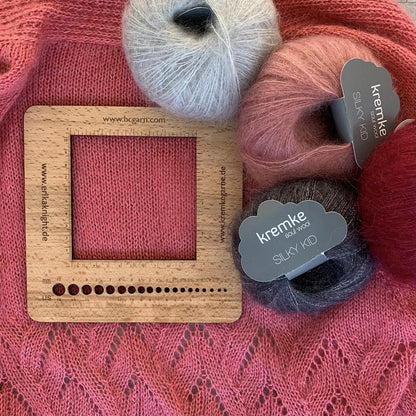 cadre en bois échantillonnier et jauge à aiguilles pour tricot sur fond de tricot rose, 4 pelotes de laines rose, grise, rouge et noire